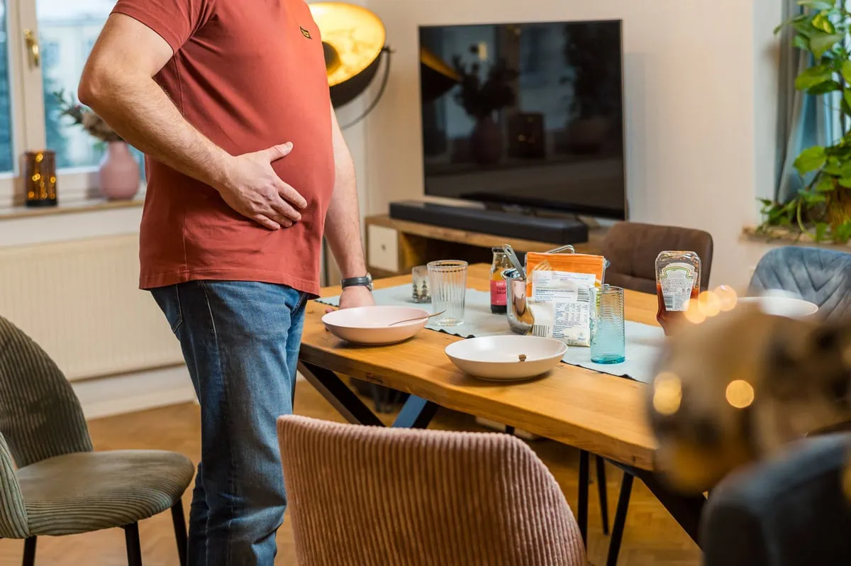Das Bild zeigt einen Mann, der sich nach dem Essen den Bauch hält und am Esstisch steht, offenbar wegen Sodbrennen.